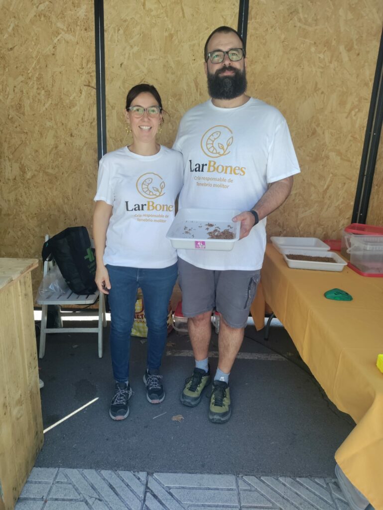 Marta i Miquel, amb samarretes de LarBones, mostrant una safata de larves de Tenebrio molitor a l'estand de FirAnoia